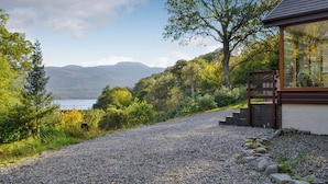 Un chemin de gravier mène à un bâtiment moderne à la façade en verre, entouré d’une végétation luxuriante et offrant une vue sur un lac et des montagnes.