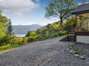 Un chemin de gravier mène à un bâtiment moderne à la façade en verre, entouré d’une végétation luxuriante et offrant une vue sur un lac et des montagnes.