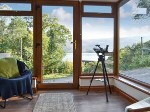 Une pièce dotée d’une grande fenêtre donnant sur un lac et des collines, un télescope posé sur un trépied, et une chaise bleue agrémentée d’un coussin vert.