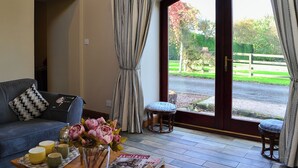 Un salón con un sofá, una mesa baja con un jarrón de flores y una revista. Hay un ventanal con cortinas y vistas a un jardín.