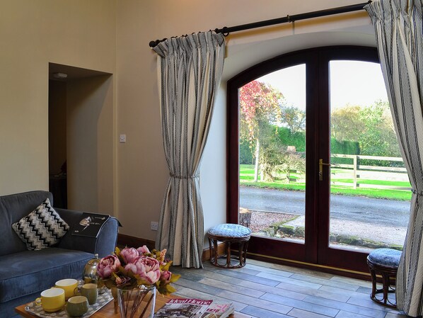 A living room with a sofa, a coffee table with a vase of flowers, and a magazine. There is a large window with curtains and a view of a garden. - 1 bedroom accommodation in Coldingham, near Eyemouth (Coldingham, near Eyemouth)