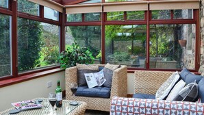 Un invernadero con mobiliario de mimbre, una mesa de vidrio con revistas y vino, y vistas a la vegetación exterior.