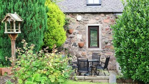 Una casa de piedra con un ventanal pequeño, un comedero para pájaros y un patio con mobiliario exterior.