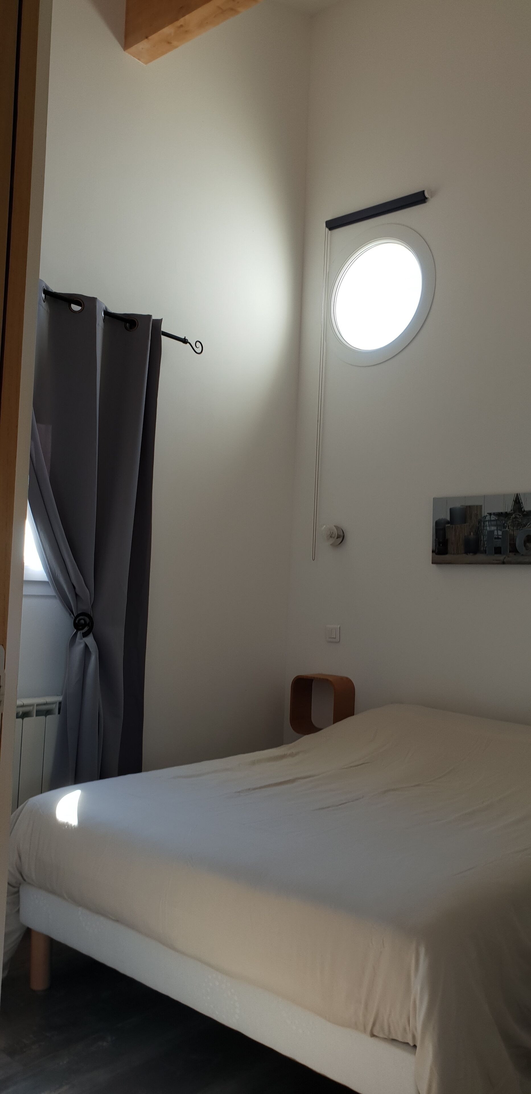 A bedroom with a bed, a small stool, a round window, and a radiator.