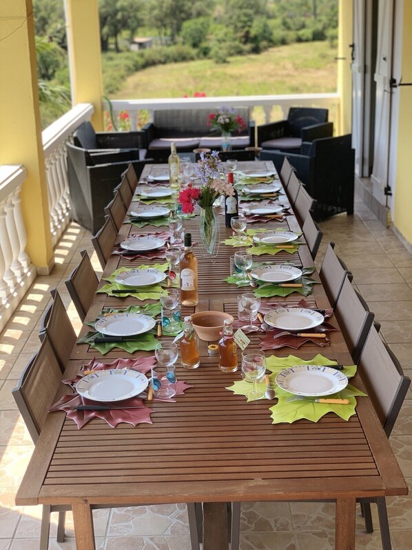 A long dining table set outdoors with chairs, plates, glasses, and a vase with flowers.