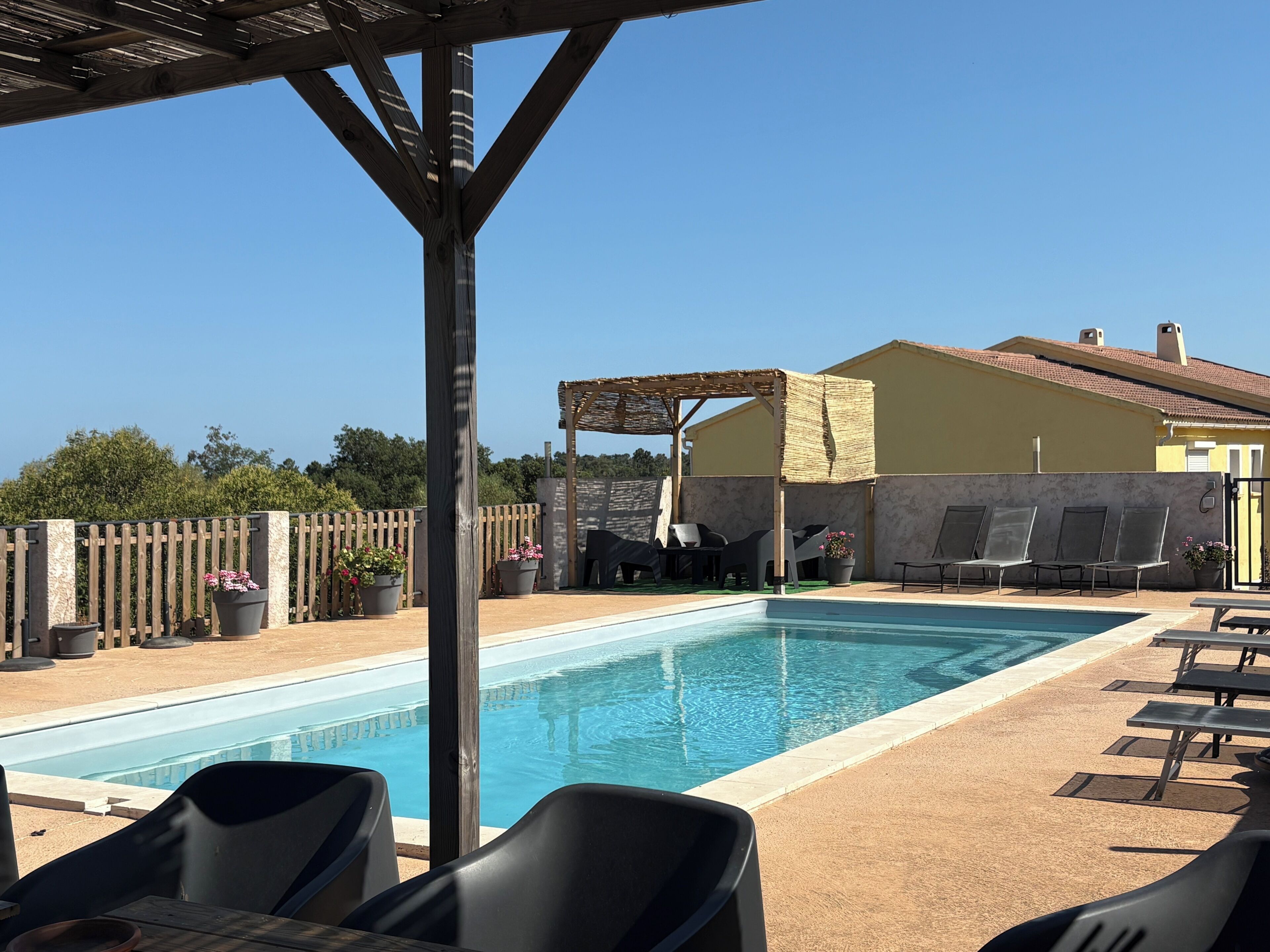 A pool area with lounge chairs, a pergola, and a building with a tiled roof.