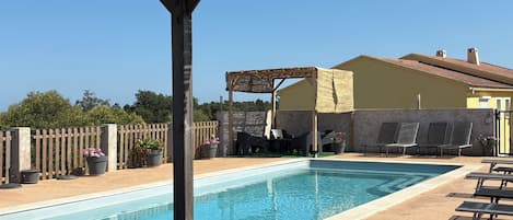 A pool area with lounge chairs, a pergola, and a building with a tiled roof.