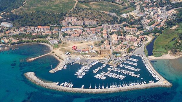 An aerial view of a marina with numerous boats docked, surrounded by a coastal town with buildings and greenery.