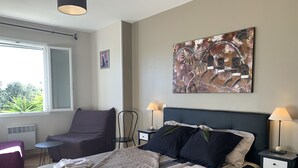 A neatly arranged bedroom with a bed, two nightstands, a chair, a small table, and a window with a view of greenery.