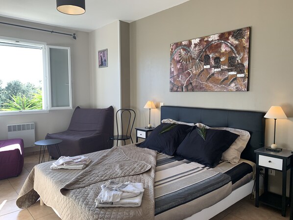 A neatly arranged bedroom with a bed, two nightstands, a chair, a small table, and a window with a view of greenery.