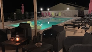 A nighttime outdoor pool area with lounge chairs and tables, a pool with floating rings, and a pergola structure.