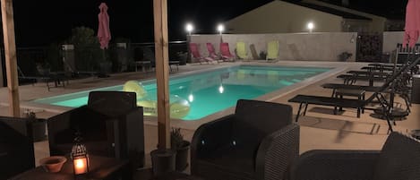 A nighttime outdoor pool area with lounge chairs and tables, a pool with floating rings, and a pergola structure.