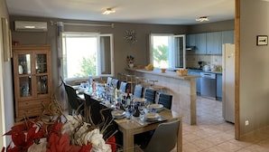 A modern kitchen with a central island, a dining area with a table set for six, and a view of the outdoors through large windows.