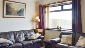 A living room with a dark leather sofa, a smaller armchair, a glass coffee table, a painting on the wall, and a window with curtains. - 3 bedroom accommodation in Uplawmoor, near Barrhead (Uplawmoor, near Barrhead)