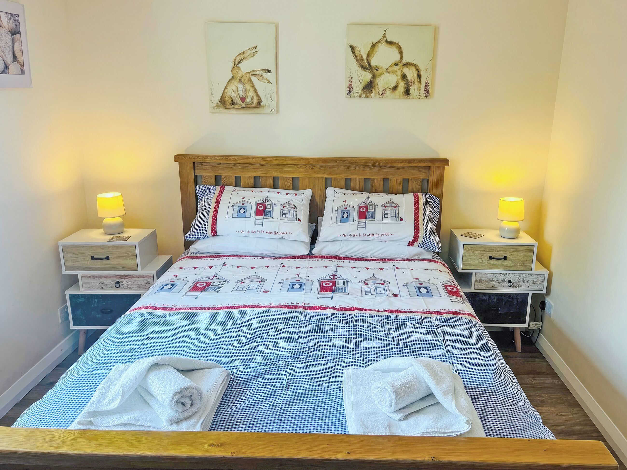 A neatly made bed with a checkered bedspread and two pillows with a lighthouse design, flanked by bedside tables with lamps and a wall-mounted artwork above.