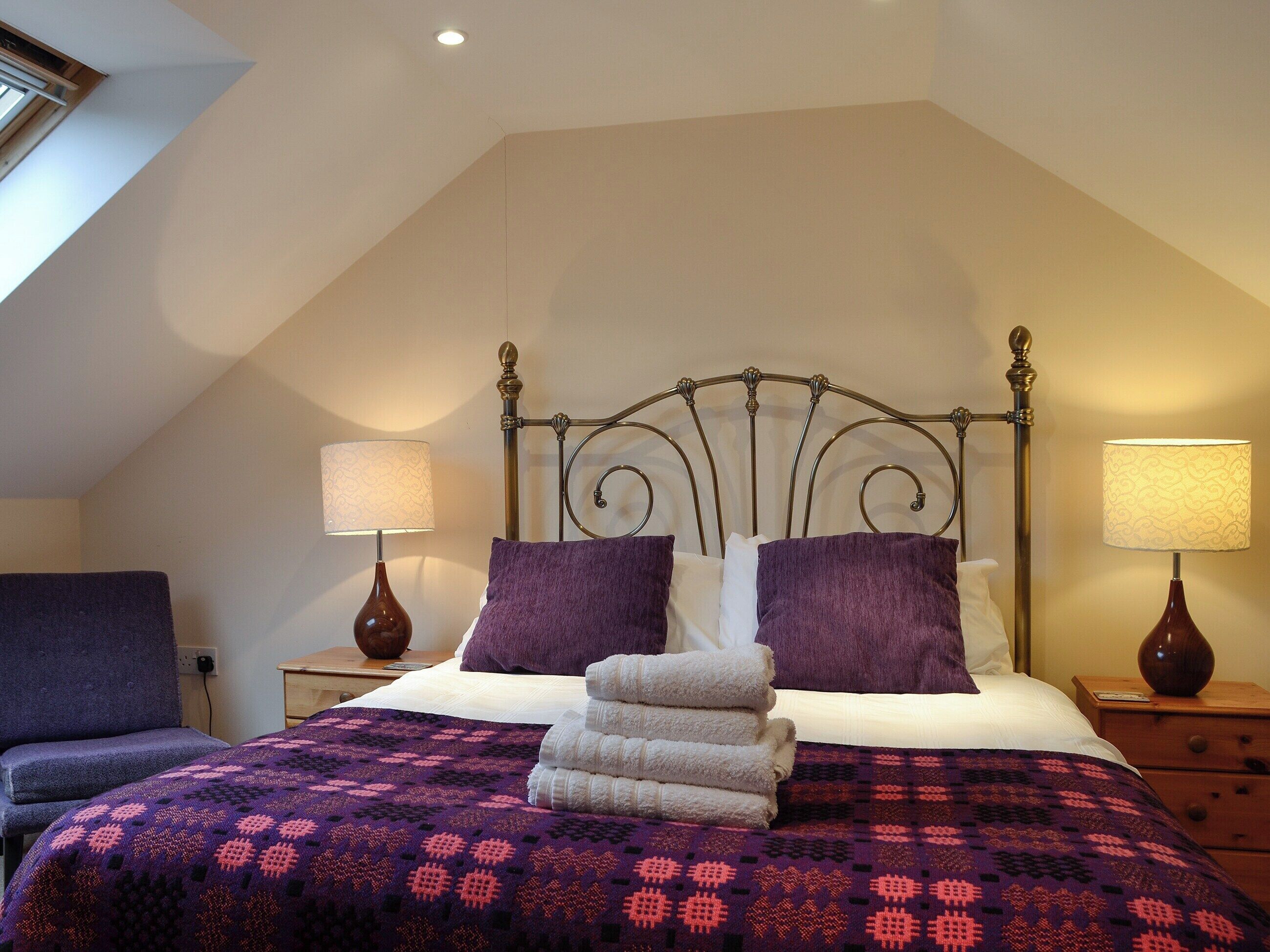 A bedroom with a sloped ceiling, a bed with a metal headboard, two bedside lamps, and a purple patterned bedspread.
