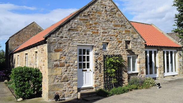 Un bâtiment en pierre avec un toit recouvert de tuiles rouges, une porte blanche et un petit jardin agrémenté d’arbustes et où se trouve un chien.