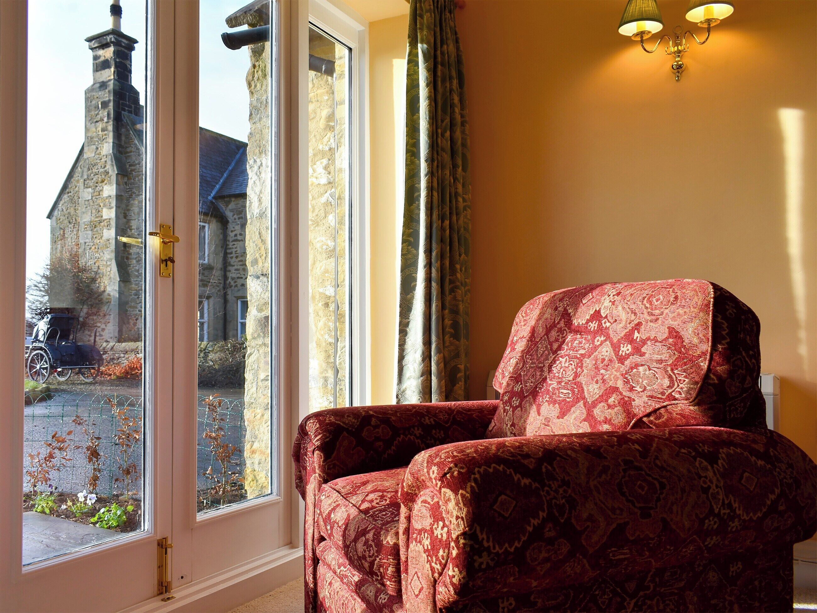 A room with a red patterned armchair, a large window with a view of a stone building and a parked car outside, and a wall-mounted light fixture.