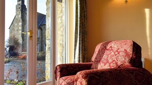 Une pièce avec un fauteuil à motifs rouges, une grande fenêtre donnant sur un bâtiment en pierre et une voiture garée à l’extérieur, ainsi qu’un luminaire fixé au mur.