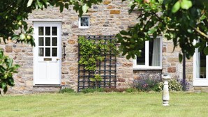 Un bâtiment en pierre avec une porte et des fenêtres blanches, entouré d’une pelouse bien entretenue et d’un petit jardin fleuri.
