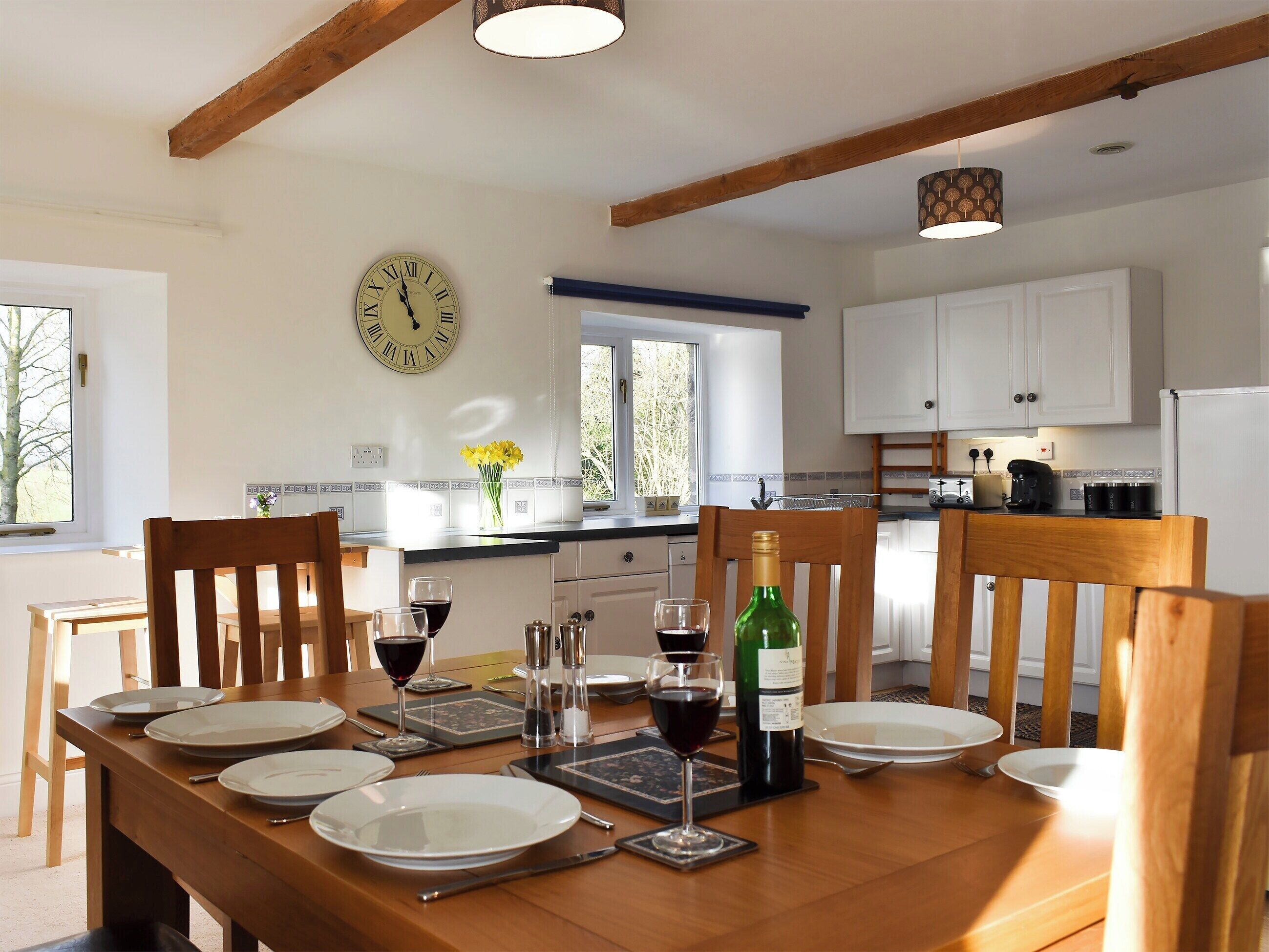 A dining room with a wooden table set for a meal, a clock on the wall, and a kitchen in the background.
