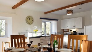 Une salle à manger avec une table en bois mise pour le repas, une horloge au mur et une cuisine en arrière-plan.