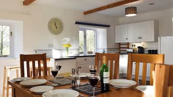 A dining room with a wooden table set for a meal, a clock on the wall, and a kitchen in the background.