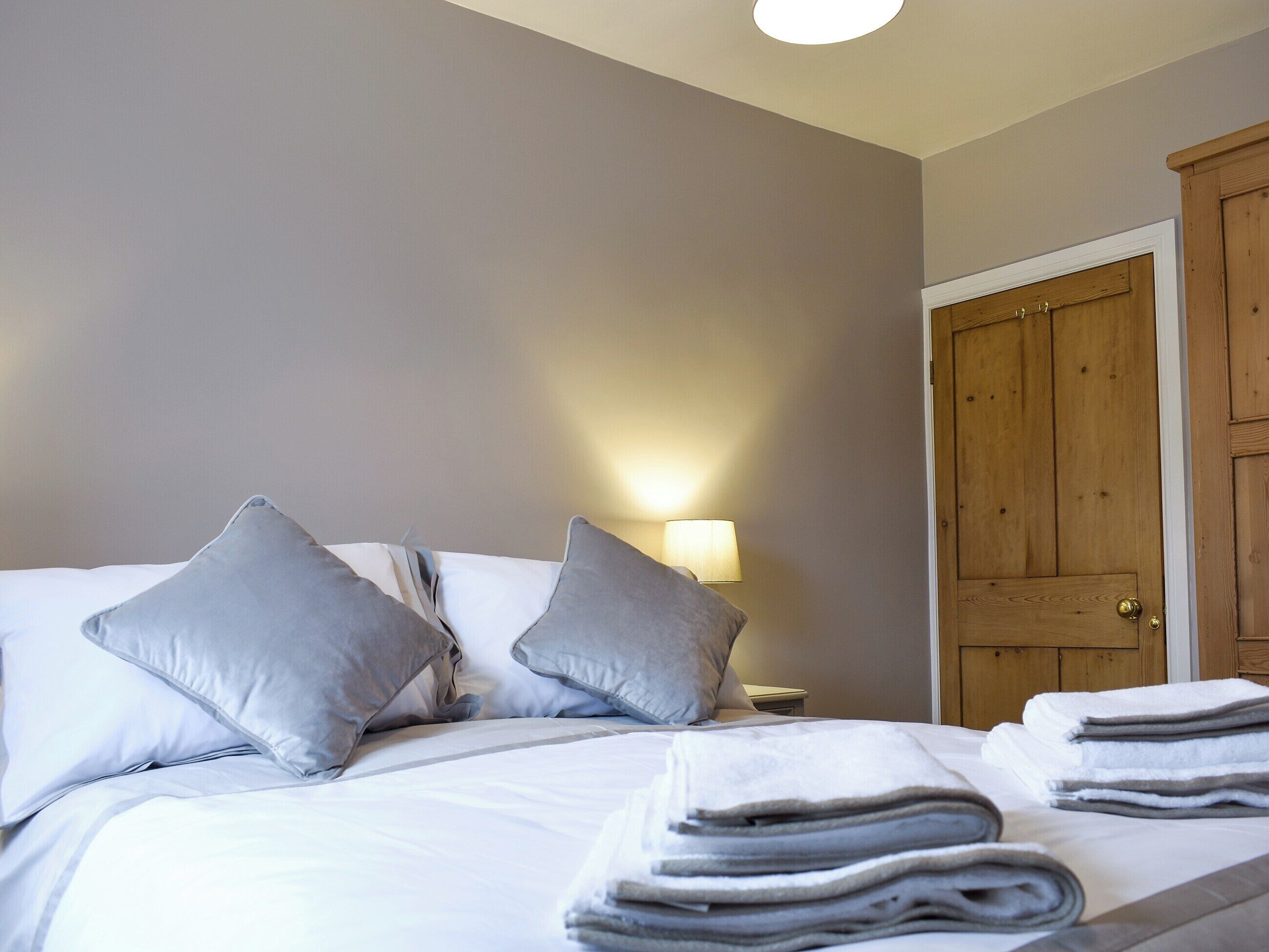 A neatly made bed with pillows and folded towels, a bedside lamp, and a wooden door in a room with plain walls.
