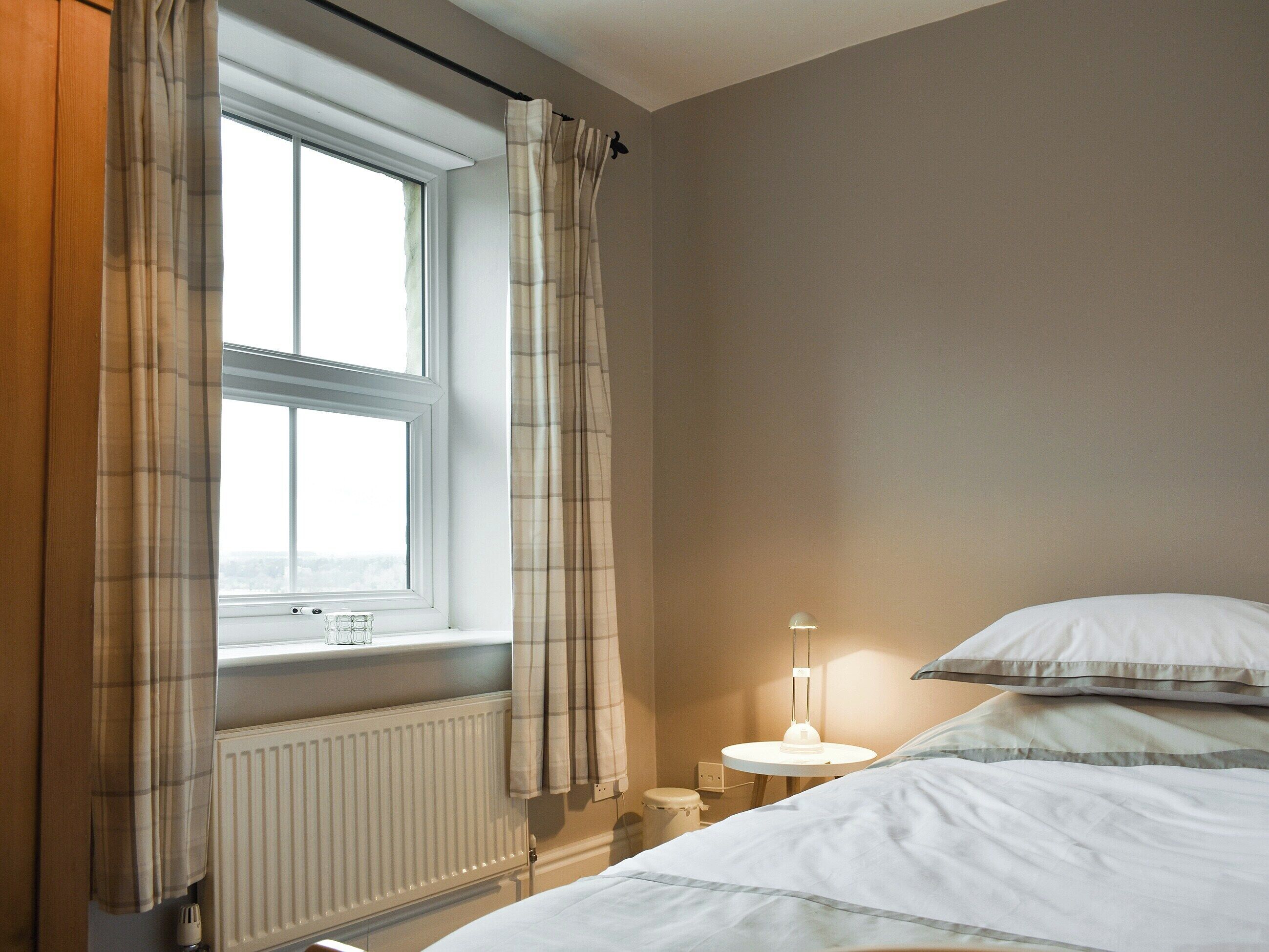 Et soveværelse med et vindue, en radiator, et sengebord med en lampe og en seng med puder.