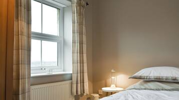 Et soveværelse med et vindue, en radiator, et sengebord med en lampe og en seng med puder.