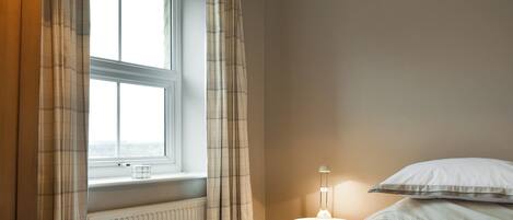 A bedroom with a window, a radiator, a bedside table with a lamp, and a bed with pillows.