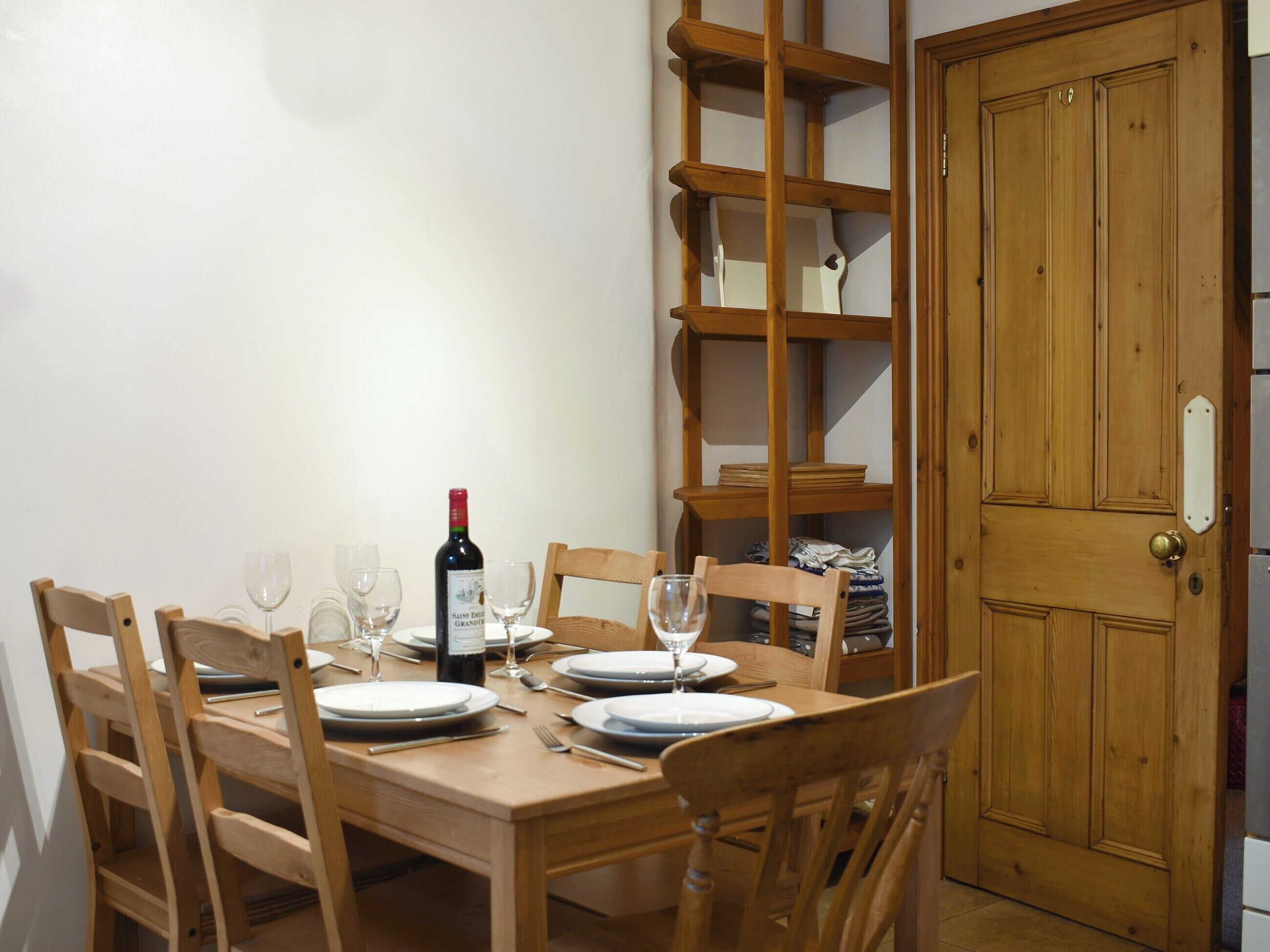 A dining table set for four with chairs, a wine bottle, and glasses. A wooden bookshelf is to the left, and a wooden door is to the right.