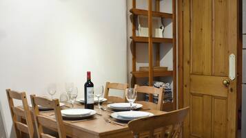 A dining table set for four with chairs, a wine bottle, and glasses. A wooden bookshelf is to the left, and a wooden door is to the right.