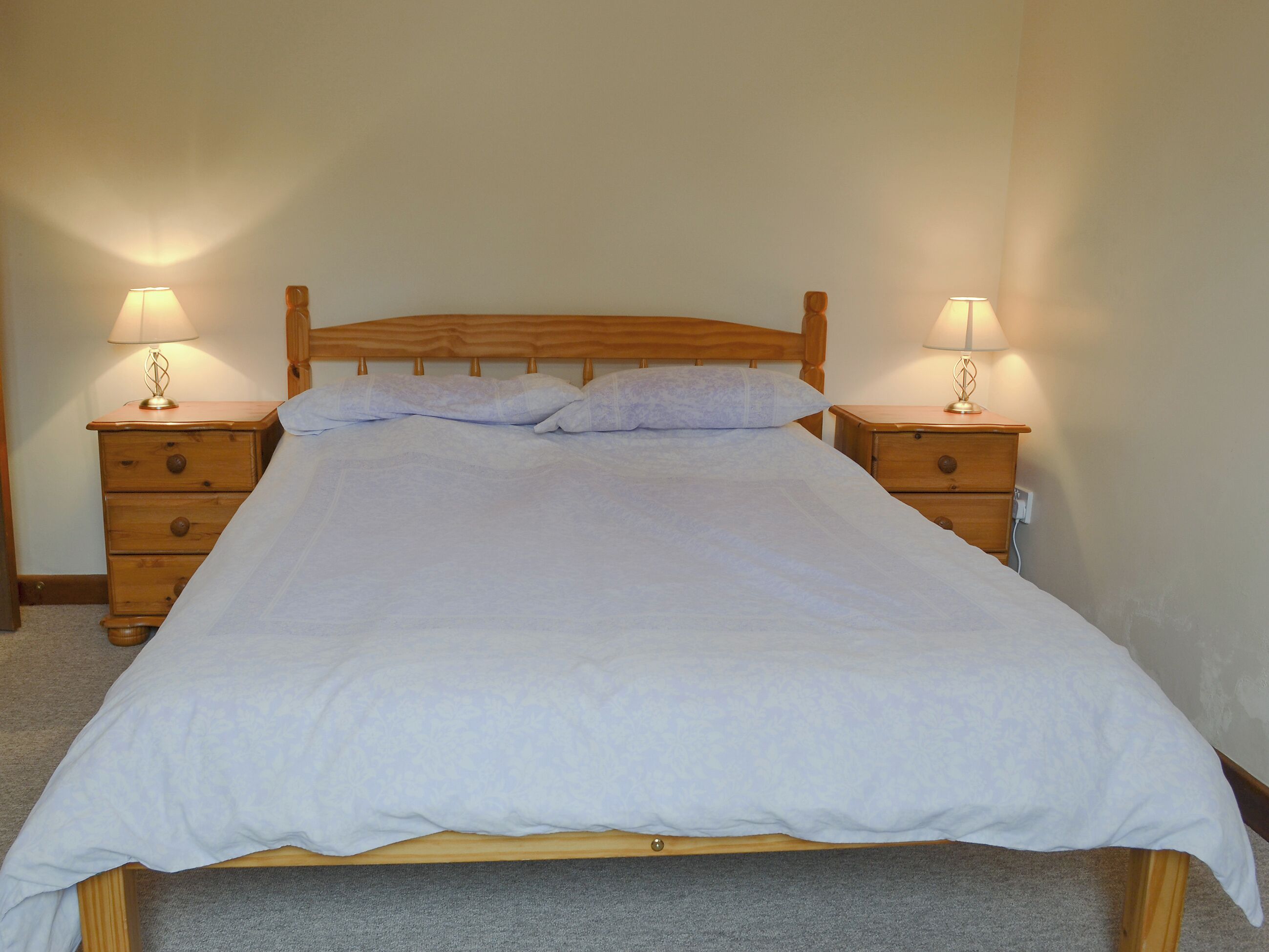 A neatly made bed with a wooden headboard, two bedside tables with lamps, and a carpeted floor.