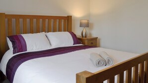 Ein ordentlich bezogenes Bett mit weißer und violetter Bettwäsche, ein Holz-Kopfteil und ein Nachttisch mit Lampe.