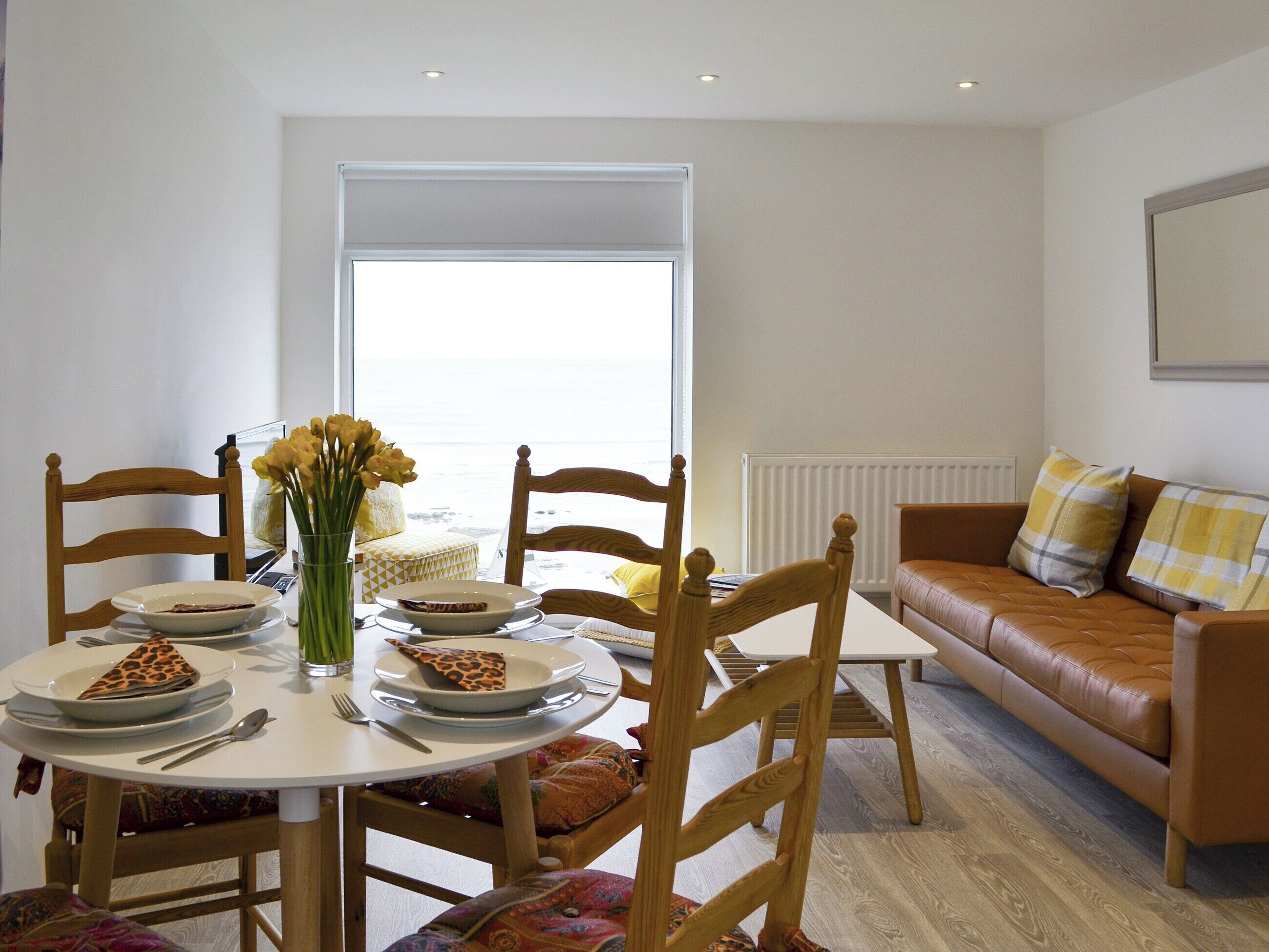 A dining area with a round table, chairs, and a vase of flowers. There is a sofa and a window in the background.