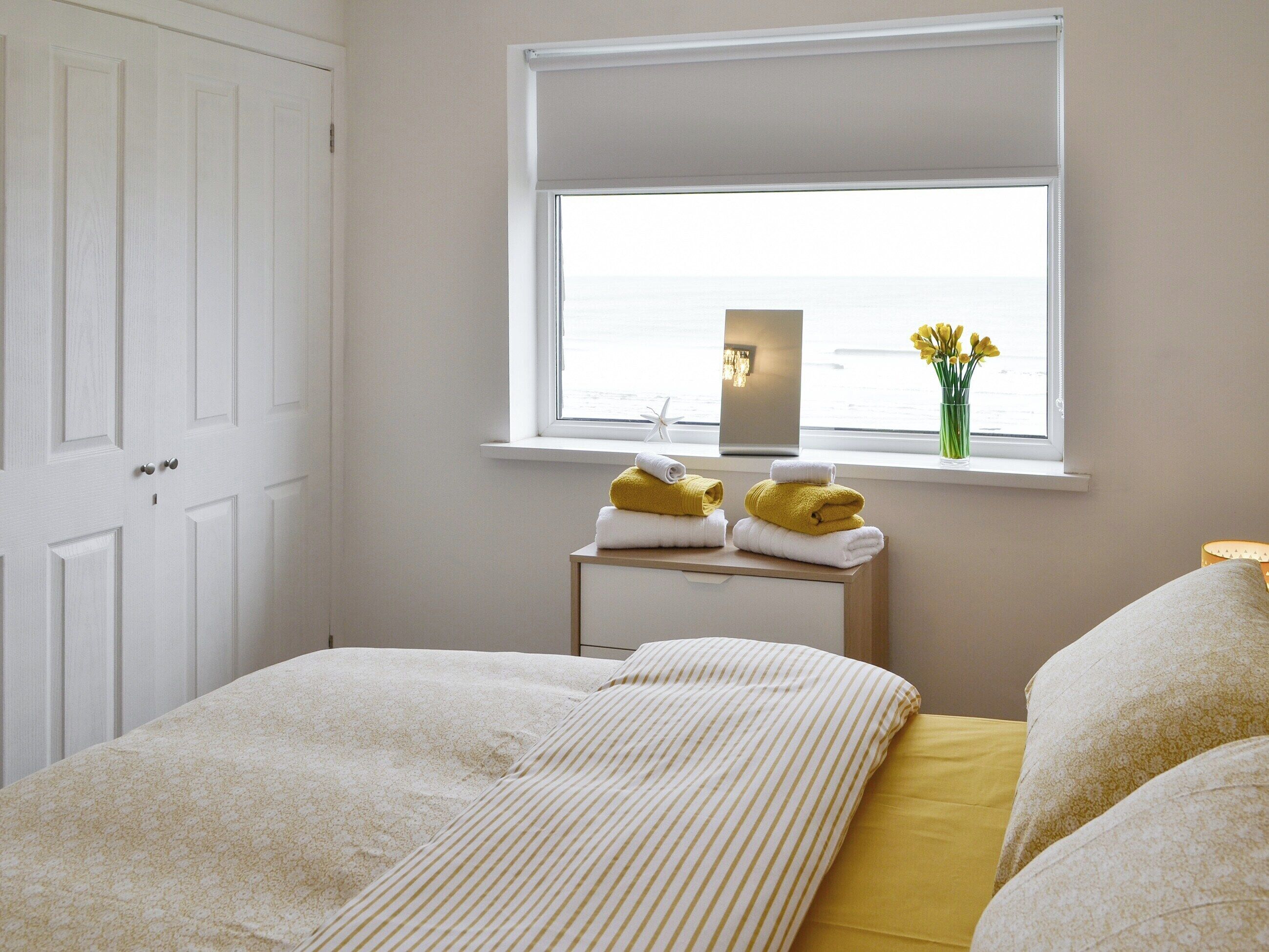 A bedroom with a bed, a window with a view of the sea, a bedside table with towels, and a nightstand with a lamp.