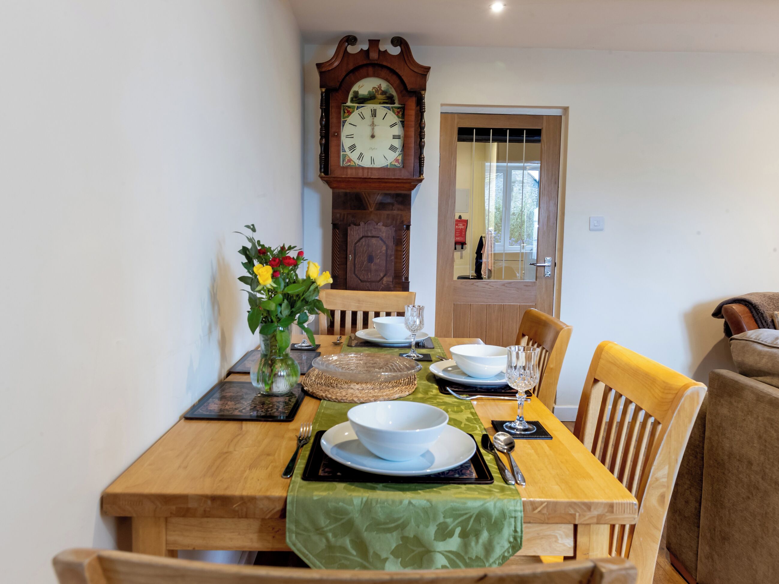 Una mesa de comedor preparada con platos, fuentes, cubiertos y un florero. Se ve un reloj de pared grande encima de la mesa.