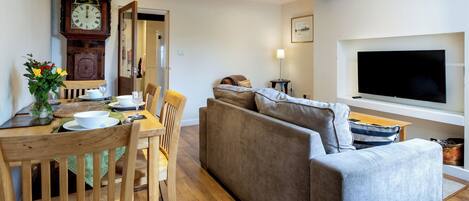 A modern living room with a wooden dining table, a sofa, a flat-screen TV, and a wall-mounted clock.