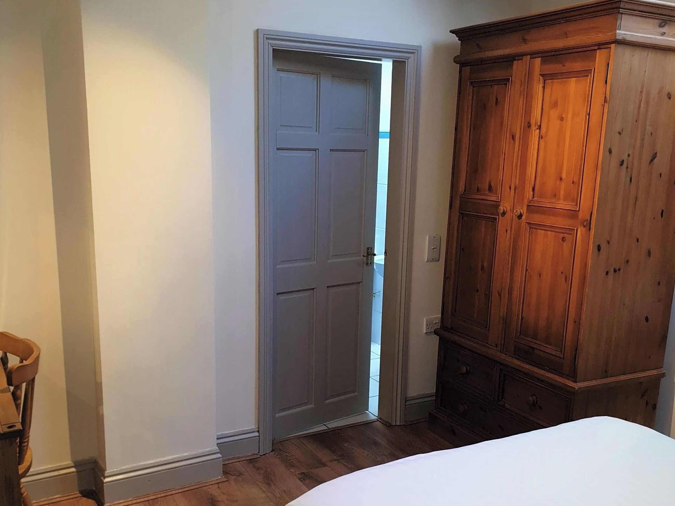 Un dormitorio con un armario de madera, una cama blanca y una puerta que da a un baño.