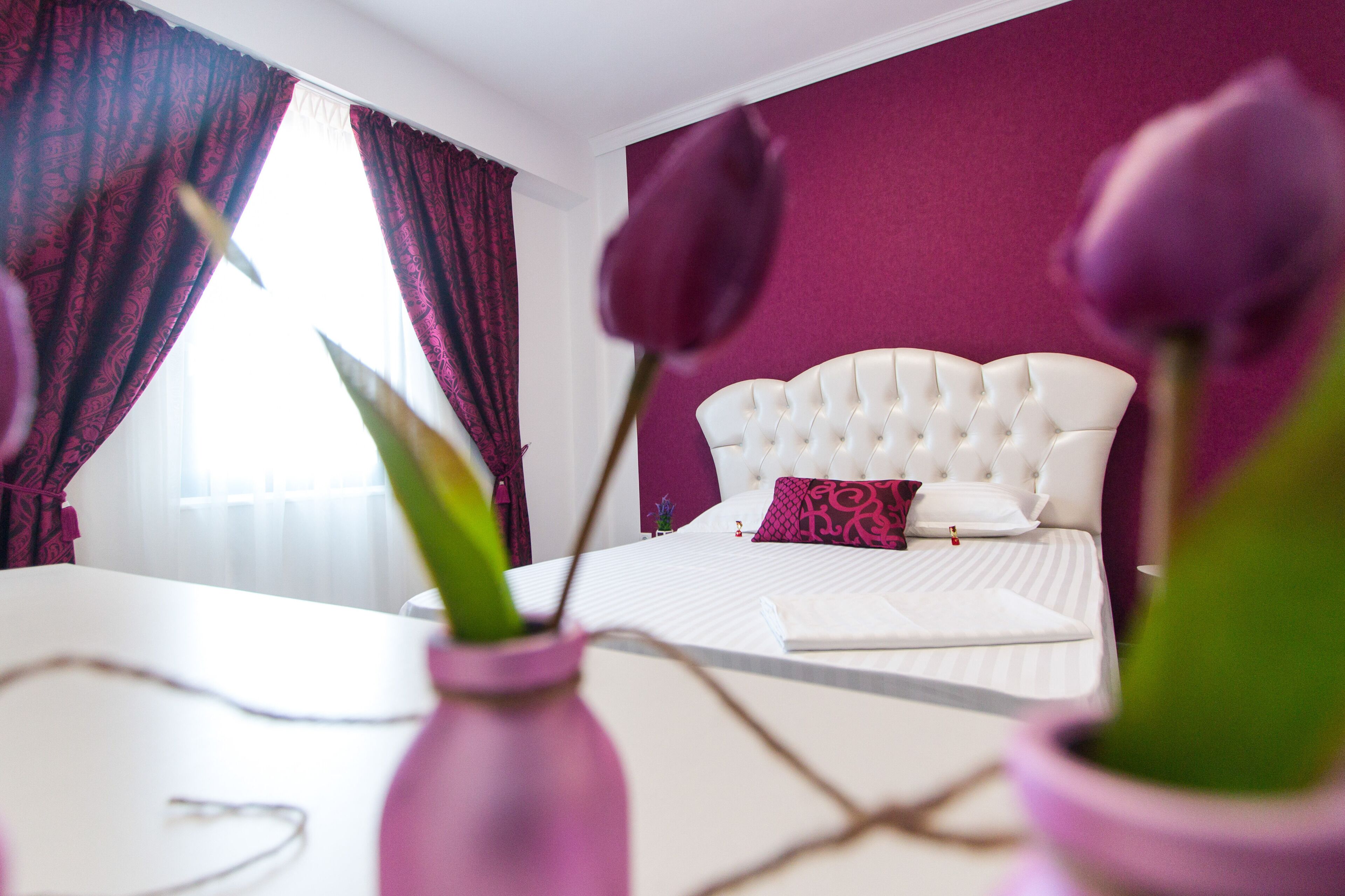 A bedroom with a white bed, purple curtains, and a vase with flowers.