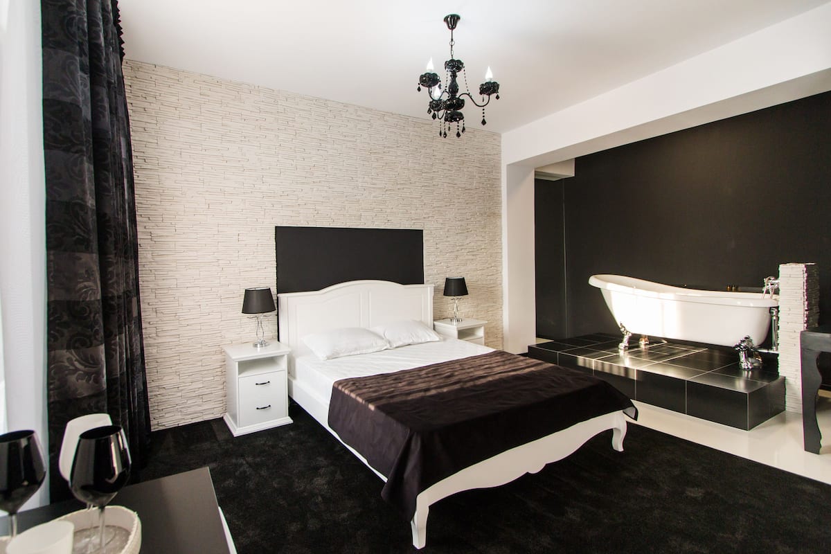 Een moderne slaapkamer met een witte baksteenmuur, een groot bed met een zwart beddegoed, een witte nachtkastje met een lamp, een zwart-wit bad en een kroonluchter.
