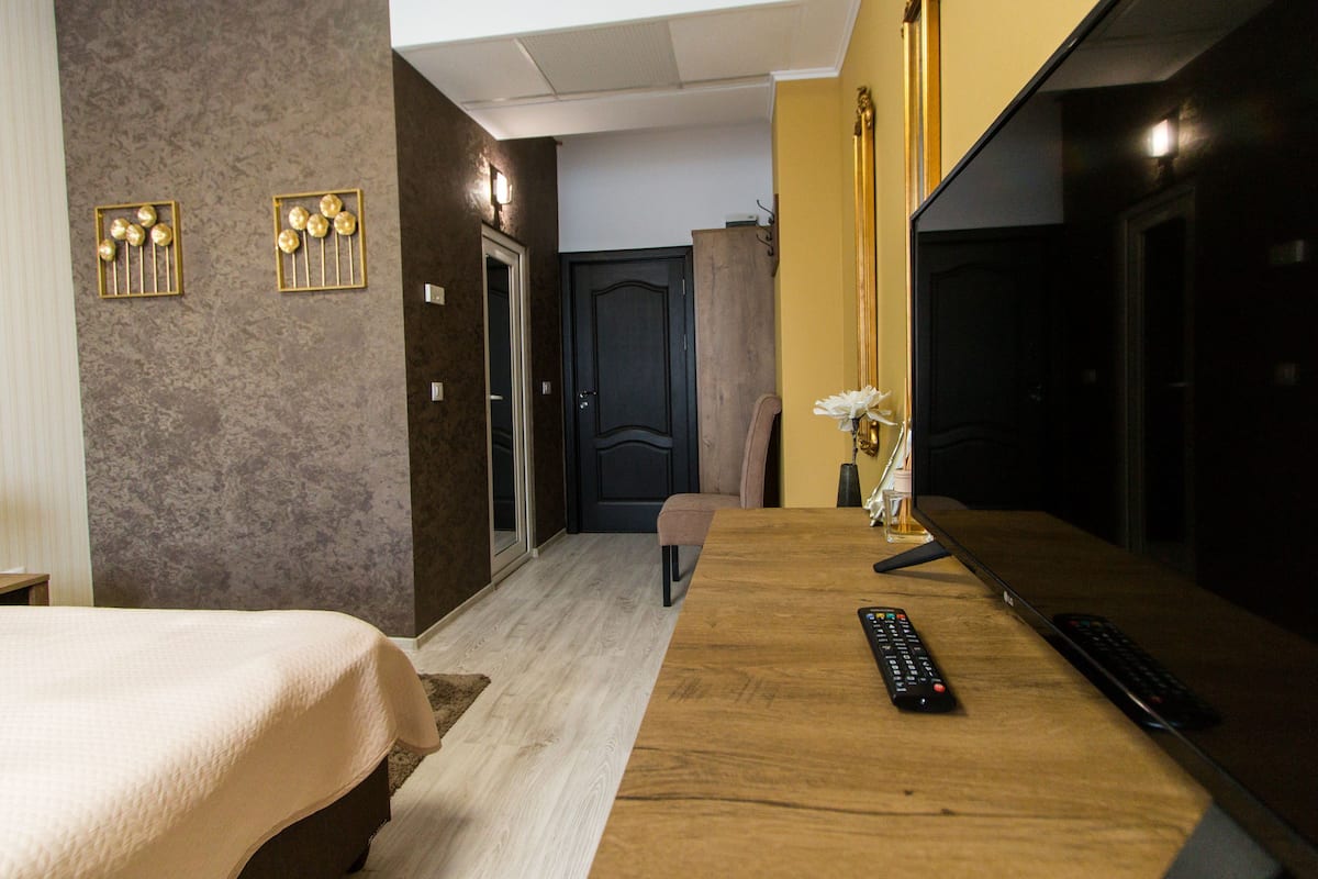 Een hotelkamer met een bed, een houten bureau, twee afstandsbedieningen, een vaas met bloemen en een deur.