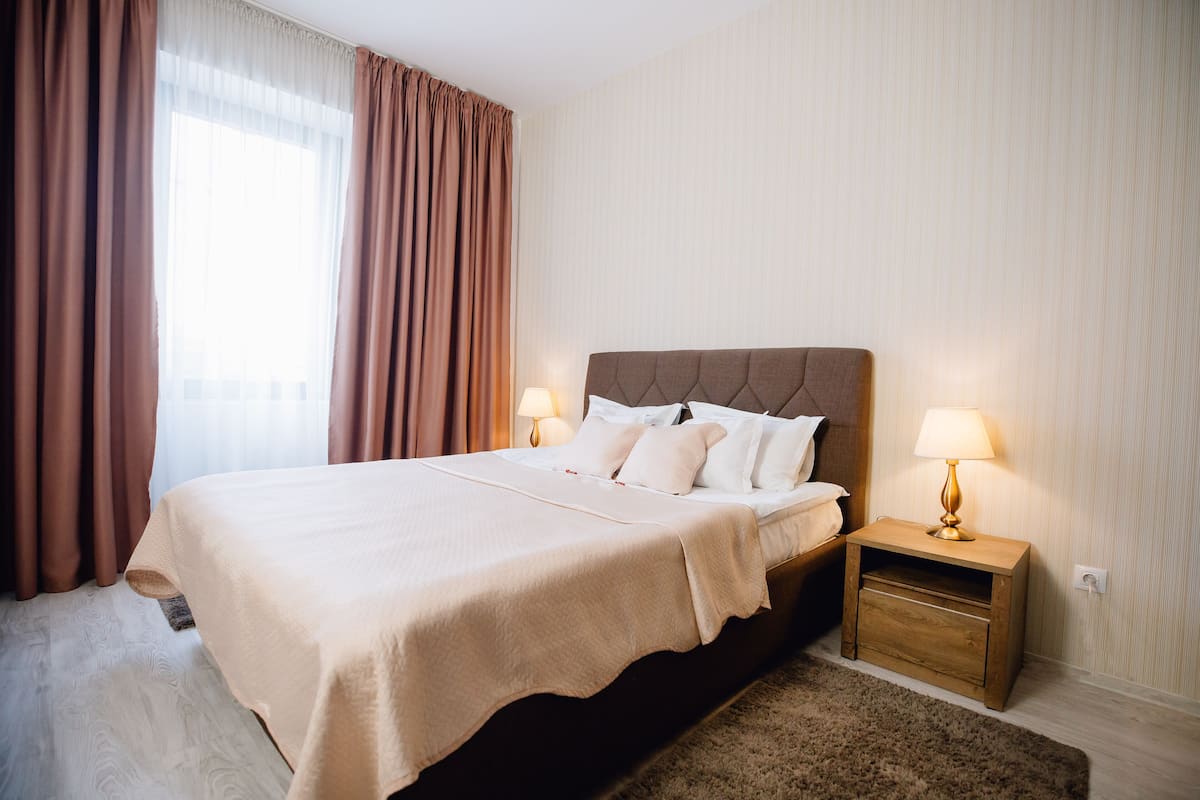 Een net opgemaakt bed met een beige sprei, twee bedlampen en een houten nachtkastje met een lamp.