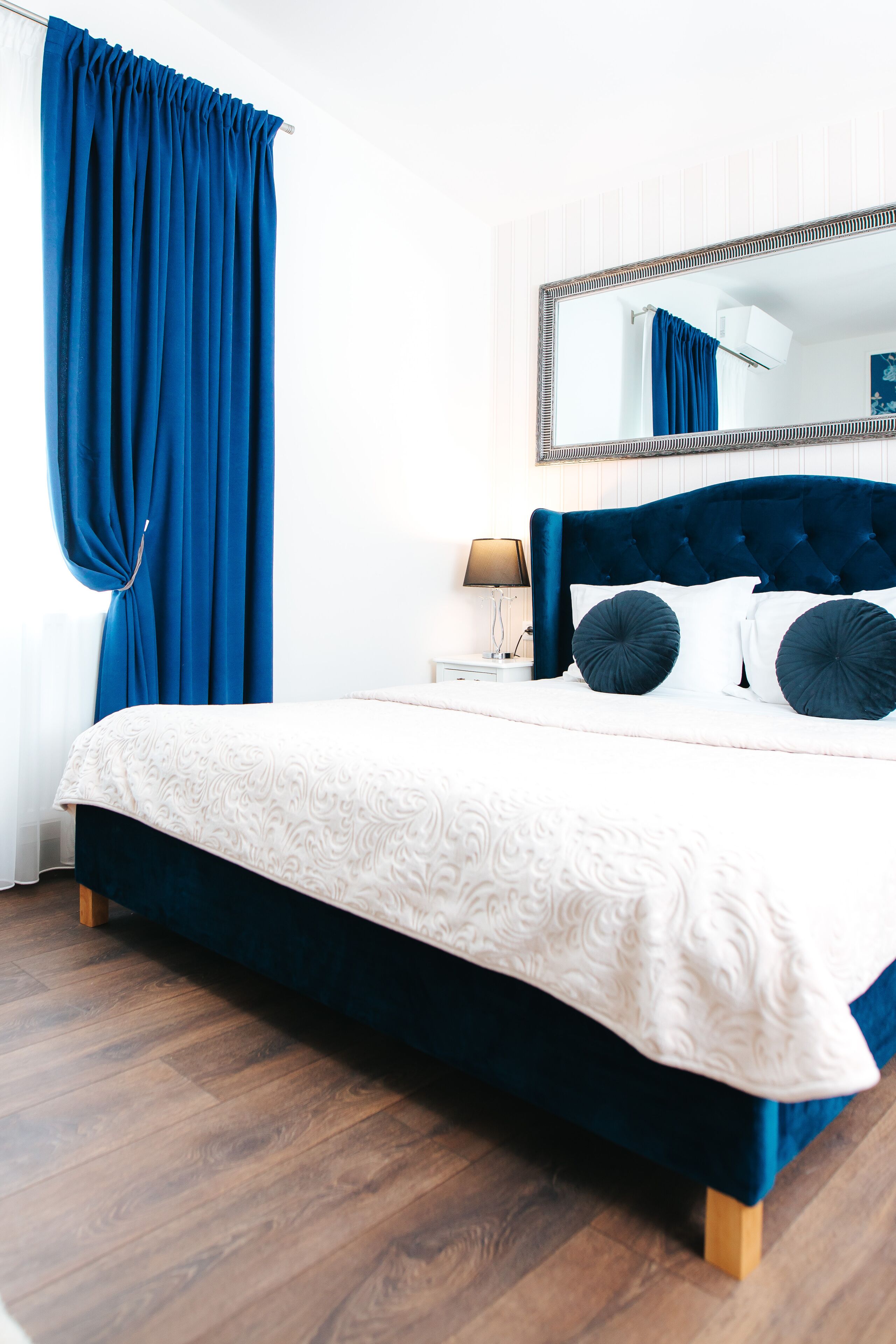 Une chambre à coucher avec un grand lit, une tête de lit, une table de chevet, une lampe et une fenêtre avec des rideaux.