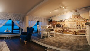 A room with a curved wall featuring a panoramic view of a cityscape, including a prominent cathedral. There are two blue armchairs, a small round table with a laptop and a bowl of fruit, and a white dining table with chairs. - Evianne Boutique Hotel (Galati)