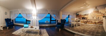 Una habitación con una pared curva que ofrece una vista panorámica de la ciudad, incluyendo una catedral destacada. Hay dos sillones azules, una pequeña mesa redonda con una laptop y un frutero, y una mesa de comedor blanca con sillas.