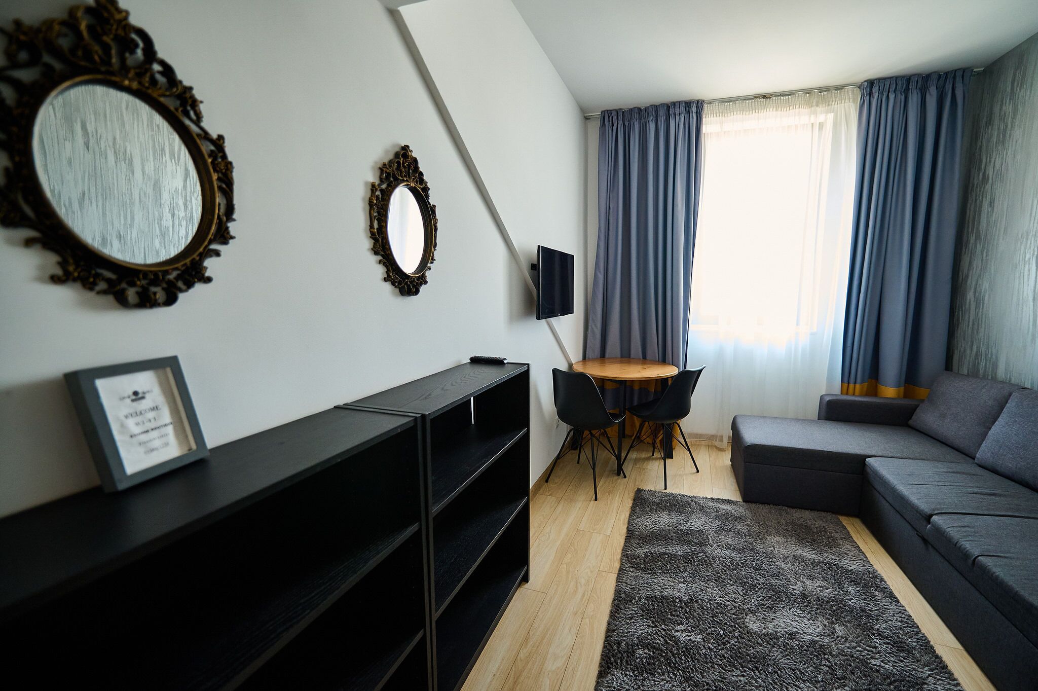 Un salon moderne comprenant un canapé d’angle, une petite table ronde avec des chaises, un téléviseur fixé au mur et un grand miroir doté d’un cadre orné.