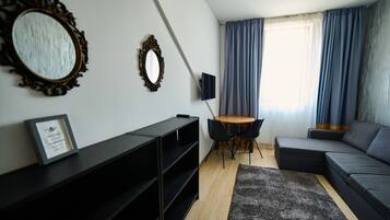 A modern living room with a sectional sofa, a small round table with chairs, a TV mounted on the wall, and a large mirror with an ornate frame.