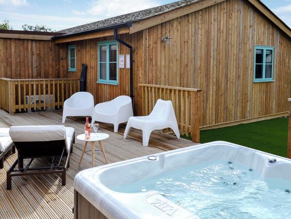 A wooden building with a hot tub, white plastic chairs, and a small table with a bottle and glass.